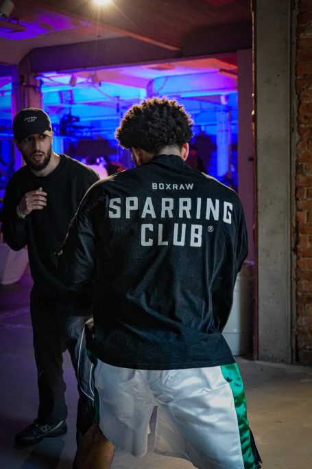 Kämpfer in Trainingskleidung mit "Sparring Club" auf dem Rücken, Training in hellem Raum.